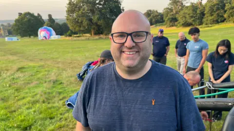 A man wearing glasses and a blue T-shirt smiles at the camera. In the background is a group of people and a hot air balloon in a field of grass.