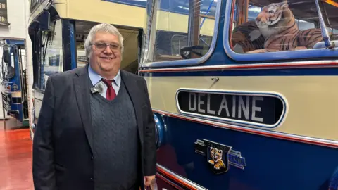 Um homem com cabelos grisalhos curtos e óculos, vestindo um blazer azul marinho, suéter de tricô, gravata vermelha e camisa azul, está parado ao lado de um ônibus vintage amarelo e azul com o texto 'Delaine' e um tigre de brinquedo no para-brisa.