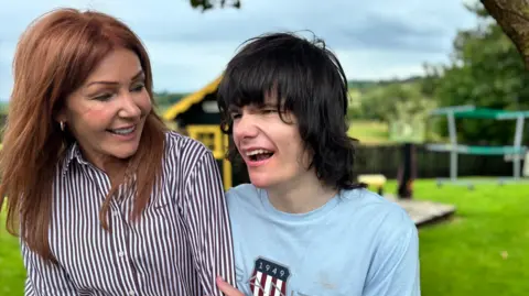 A mother and her son sit in a bright garden. The woman is smiling down fondly at her son as he holds her arm. She has long red hair that is being blown by the wind and is wearing a shirt with stripes. The young man alongside her has a big smile. 