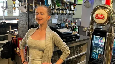Jonny Humphries/BBC Antonia Merner, with blonde hair tied back in a bun and a grey cardigan over a grey top, smiles at the camera behind the bar.