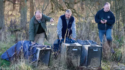 Chiew Loo Three men, standing by three cages, the man on the left, is holding up a cage door, wearing a green coat, light trousers and dark top, the man in the middle is resting his hands on a cage, and has on a blue shirt, blue gilet and has a beard. A man is standing to the right, wearing jeans, boots and a black hoodie. 