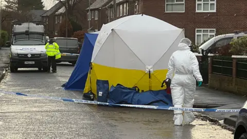 A road with blue and white police tape with a forensic investigator walking towards a white and yellow tent in which has been erected within the cordon