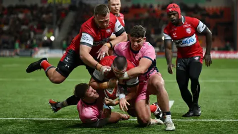 Getty Images A group of players commit to a tackle as Gloucester play Exeter in the Premiership Rugby Cup at Kingsholm. A Gloucester player is going to ground with the ball in his hands, while two Exeter players tackle him, one of them grimacing with the effort. In the background other Gloucester players look on. Gloucester are in their traditional colours of red and white while the Exeter players are in their change kit of light pink and black