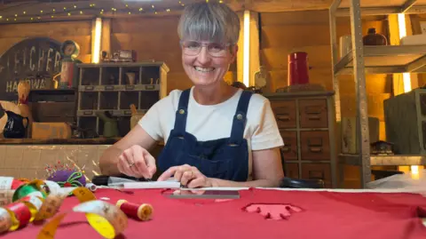 Rebecca Bissonnet, with short grey hair and wearing black dungarees over a white t-shirt, is restoring the red vintage football shirt.