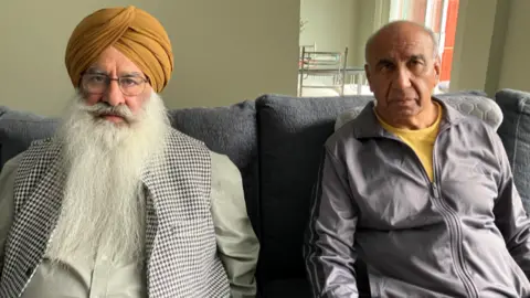 Mr Singh has an orange turban and a long white beard and glasses. He is wearing a light coloured shirt with a checked waistcoat. Mr Sangha has a yellow T'shirt on and grey tracksuit top on. He has short grey hair.
