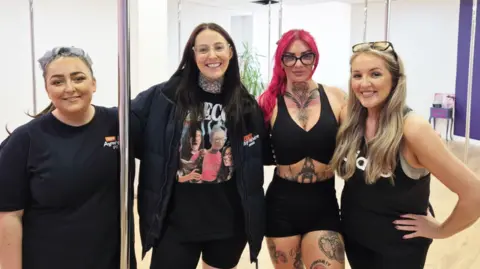 BBC Four women, standing in a dance studio, with several silver poles visible behind them or next to them. The group have one woman wearing all back, with dark hair to her shoulders, another woman with glasses, a dark T-shirt and jacket on and long, dark hair past her shoulders, a third woman with dyed red hair, a black crop top and black shirt, and the last woman has blonde hair, glasses resting on the top of her head and is wearing black workout gear.