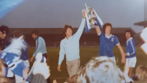 PAT EDWARDS/PHOTOEAST Ipswich Town footballers show off their FA Cup winning trophy to fans at Portman Road in 1978. Two players hold the trophy above their heads. One wears an Ipswich Town kit and the other wears a blue jumper with chino trousers. 