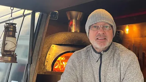 Mark Clarey with a grey beard wearing silver glasses a grey beanie hat and grey fleece inside his pizza van with the flames from the pizza oven in the background. A lantern hangs to his left from the hatch door. He is smiling. 
