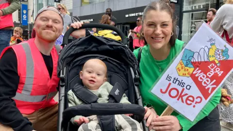 Nicola Rees/BBC A man and woman kneeling next to a pram with a baby in it on a busy high street, all smiling at the camera.