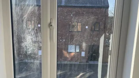 Tyllar Mellor The sun is shining on a window which is badly scratched. You can see a house behind the window.