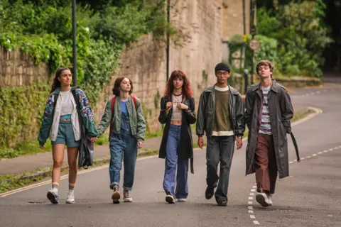 BBC/Moonage Pictures A group of five young people, three women and two men, are walking in line on the street, with two of the girls are holding hands