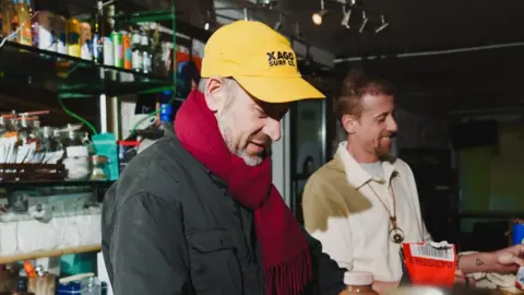 ARC Two men are making drinks at the bar. Javier Tanke is wearing a dark jacket, red scarf and yellow baseball cap, while the other man in the background is wearing a cream fleece. 