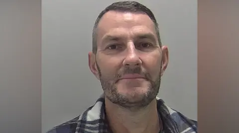 Cambridgeshire Police A man with dark hair and a beard in a blue and white checked shirt