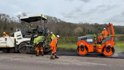 A road with obvious cracks is being laid with fresh tarmac by a large white machine. It is being rolled over by a large bright orange roller, driven by a man in orange high visibility clothing. There are several workmen wearing high visibility clothing and hard hats, sweeping the tarmac. There is a green grass verge and a hedgerow next to the road.