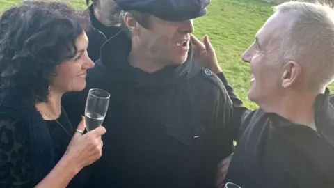 Alison Bell Alison Bell, wearing black and holding a champagne flute, and Liam Walsh, wearing black, smile and laugh as they speak with Liam Gallagher, who is wearing a black cap and black coat