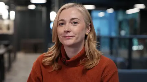 Foto kepala dan bahu Jess Blair, yang mengenakan jumper merah dan duduk di kantor BBC di Cardiff.