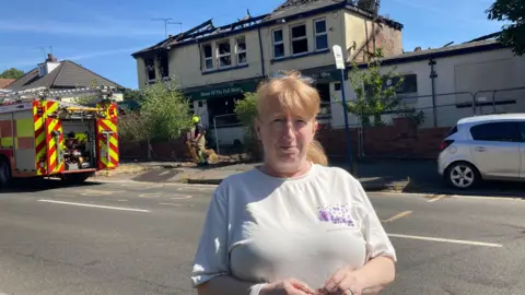 BBC/Naj Modak Teresa Hodges is in a white T-shirt standing across the street from a fire-damaged building surrounded by temporary fencing. A fire engine is parked on the left, a white car is parked on the right, and the building's damaged roof and broken windows are visible in bright sunlight.
