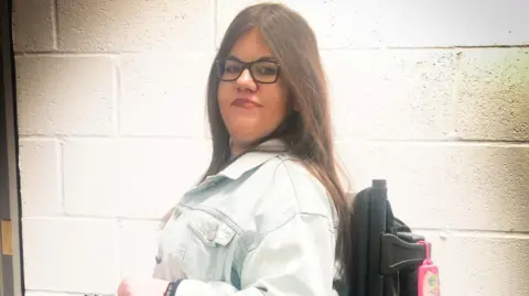 Sara Stacey smiling straight at the camera. She is wearing a denim jacket and black glasses and is in front of a white brick wall.
