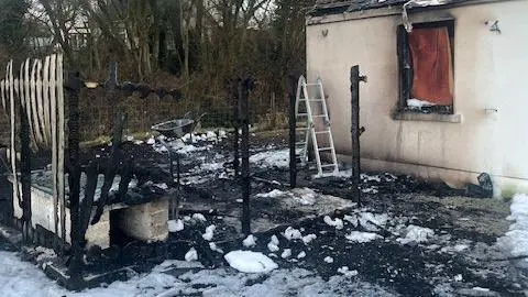 Victoria Nichols The back of the house showing where the oil tank once stood - it is surrounded by the remains of a wooden fence which has been burnt down, only blackened ashes remain. On the ground ashes and foam from the firefighting can be seen. An outbuilding is to the right of the image.