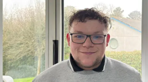 Dylan Caulkin, a young man with short curly hair in glasses, smiling wearing grey sweatshirt and collared T-shirt, standing in front of a window looking out on a hedge and tree