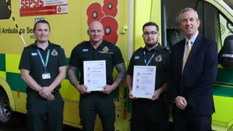 EEAST Three people are in ambulance uniform and one is in a suit. They are stood in front of an ambulance which has a poppy design on the side. The two people in the middle are holding white certificates. 