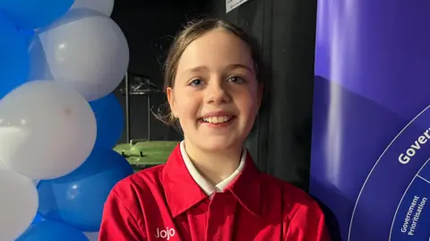 An 10 year old girl with brown hair tied back, wearing a red boiler suit with the words Team Leader below her name, Jojo. She is looking straight down the camera and smiling. She has blue and white balloons to one side of her and a purple banner to the other side. 