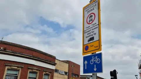 A sign which lists the street is a pedestrian cycle zone, and only buses and loading vehicles can use it. A sign stating cyclists can use the road too is on the post.