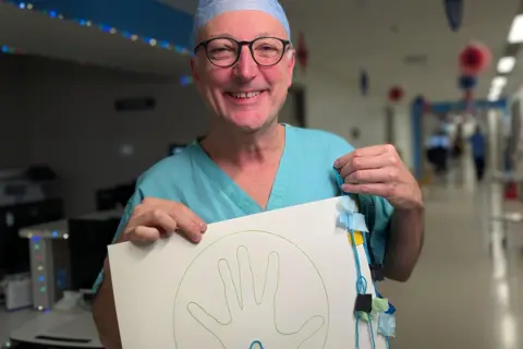 Consultant Dr Giampaolo Martinelli, a male consultant in scrubs in a hospital corridor, holding his artwork 