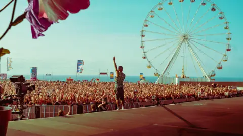 A performer stands on a stage facing a large crowd at an outdoor festival by the sea, with many people raising their hands. In the background, a large Ferris wheel rises above the crowd under a bright blue sky, while colourful flags and decorations add to the lively atmosphere.