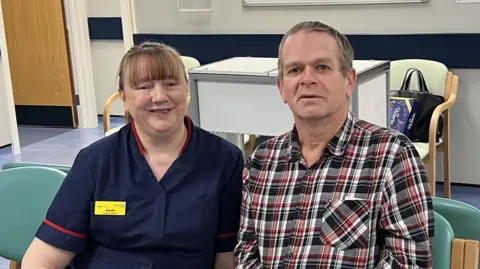 Kettering General Hospital Ophthalmology Matron Sarah Robinson sits beside Michael Knighton and they both smile at the camera. They are sitting in a hospital waiting area, Ms Robinson is wearing a navy nurses uniform and Mr Knighton is wearing a plaid shirt.