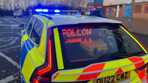A general view picture of a police car with a light which says, in red lettering, "follow" illuminated in its boot.