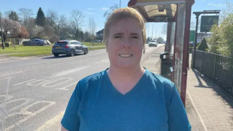 BBC/Seb Cheer A woman with short blonde hair stands near a bus stop, smiling at the camera. She is wearing a blue top.