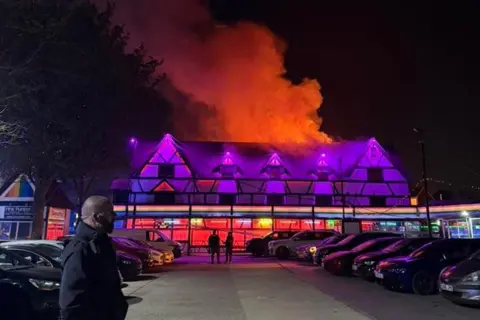 Gabby Cantareli The outside of Pink Punters, a night club in Fenny Stratford. There are purple neon lights and smoke and orange flames coming from the top of the building.