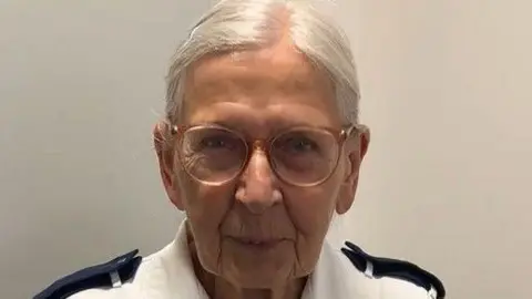 Iva smiles directly into the camera.  Her eyes are twinkling behind her clear light pink glasses and she wears a white nurses uniform.