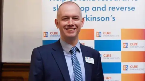 Cure Parkinson's Simon has very short hair and is wearing a navy suit jacket, blue shirt and blue and white tie. He is standing in front of a blue and orange banner with the Cure Parkinson's logo on it