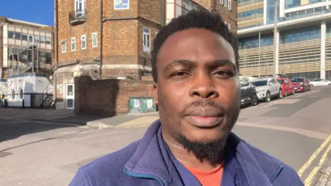 Chidi Ezikpe, wearing a purple fleece and orange t-shirt, stands in a street in Brighton.