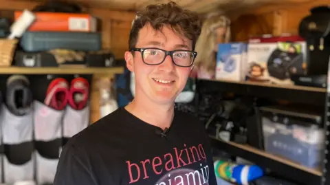 Alex Dunlop/BBC Matt smiles at the camera. He is wearing black-rimmed glasses, has brown, short, wavy hair and a black T-shirt with a graphic. He is standing in a shed filled with shelves and goods. 