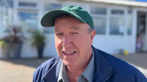 A man wearing a green baseball cap, blue zip up jacket and striped shirt smiles for a photograph. 