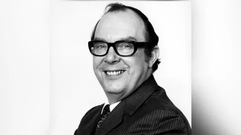 Black and white photo of Eric Morecambe who wears a suit, tie and glasses and stands in a side profile stance
