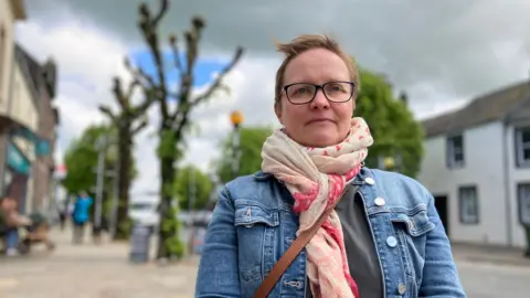 Trella Javanainen standing on Main Street in Cockermouth. She has short strawberry blond hair and is wearing glasses with a dark rim, a denim jacket, pink pashmina and grey top. She is looking serious towards the camera. The background is blurred and there is a pollarded tree behind her and shops.