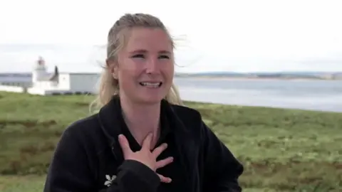 Sophia Jackson, who has blonde hair, holds her manus  to her thorax  looking happy. A achromatic  lighthouse is successful  background. 