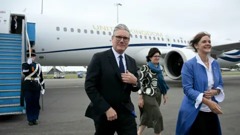 PA Media Sir Keir Starmer arrives at Amsterdam airport ahead of a Nato summit. There are Dutch soldiers forming a guard of honour. Sir Keir is wearing a dark suit and dark blue tie.