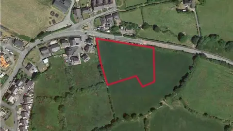 Cyngor Gwynedd Plans Aerial drone shot shows the proposed area for the housing estate. A red line shows the area on a green field next to a village. 