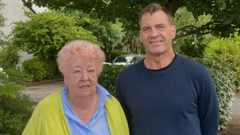 Two people standing looking into the camera. Woman has pink hair and a green cardigan and a blue top. Man has a navy blue jumper on. 