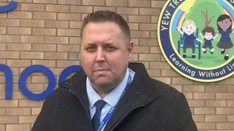 Jamie Barry is stood in front of the Yew Tree Primary School sign and brick building. He is wearing a chequered shirt and blue tie with a black coat on.