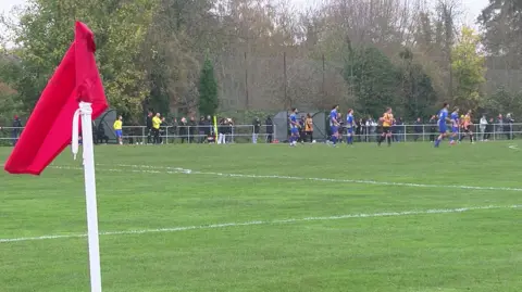 BBC A scene from football being played at Faraday Road in Newbury, where Newbury FC (playing in orange and black) are playing against a team in a blue kit. A red corner flag is pictured on the left side of the picture, with a handful of spectators lining the pitch