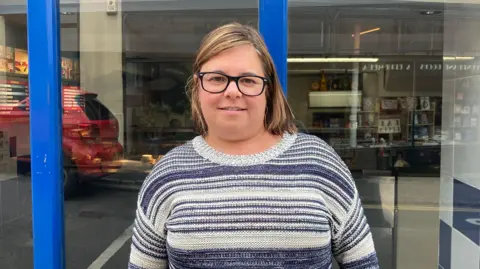 A photo showing a white woman stood in front of a shop window. She has brown hair and is wearing glasses and a blue and grey striped jumper