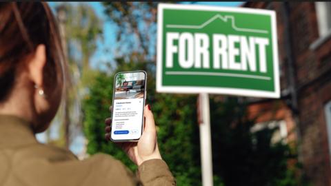 Women looks at mobile phone in front of For Rent sign