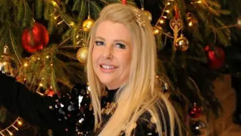 North West Anglia Hospitals' Charity Natalie Styles-Hudson is in the middle. She has long blonde hair and is wearing a gold clip on the right side. She is wearing a black sequin top and is in front of a Christmas tree with gold and red baubles. 