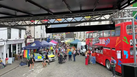 Ulverston Pride Festival The photo, taken from a stage, looks out onto multiple brightly coloured stalls. There look to be about 100 people.